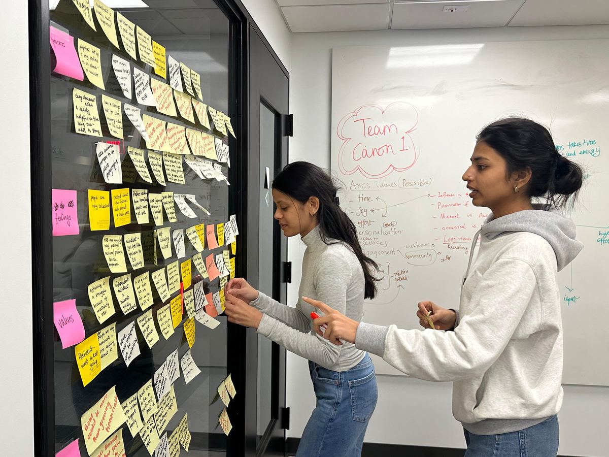 Two teammates clustering sticky notes on a glass wall beside a whiteboard headed Team Canon 1 with value axes